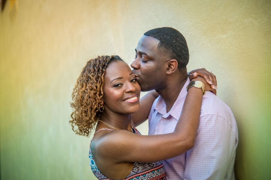 Engaged man kissing his fiancée on the cheek as she smiles against a warm, softly lit wall