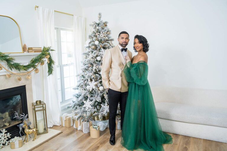 Couple in an emerald tulle gown and champagne tuxedo jacket beside a white flocked Christmas tree