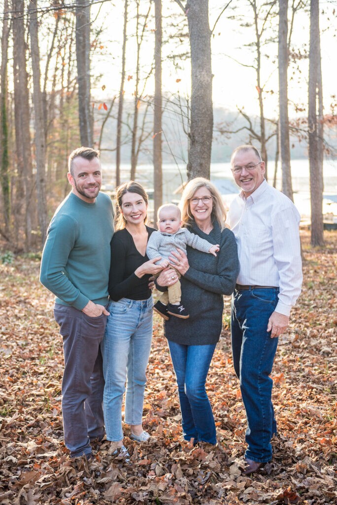 Three-generation family standing together in fall leaves by a lake, wearing coordinated fall family photo outfits in teal, black, white, gray, and denim