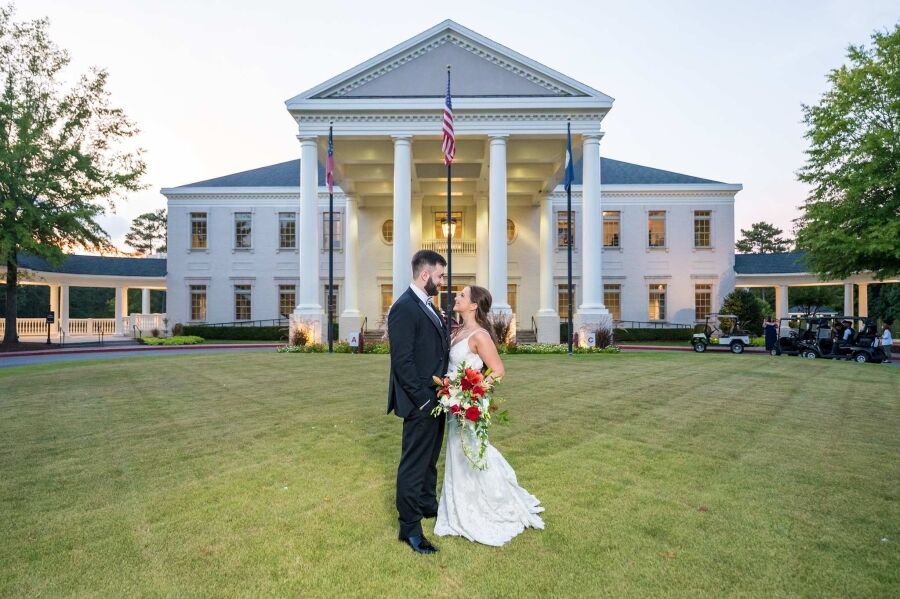 Bride and groom stand centered on the lawn in front of the columned Governors Towne Club clubhouse
